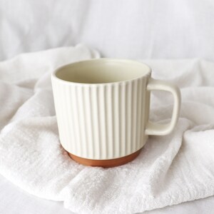 Può includere: Una tazza in ceramica bianca con un design a coste e una base marrone. La tazza ha un manico ed è appoggiata su una superficie di tessuto bianco.