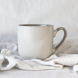Puede incluir: Una taza de cerámica blanca con un asa de color marrón claro. La taza está sobre un paño de lino blanco.