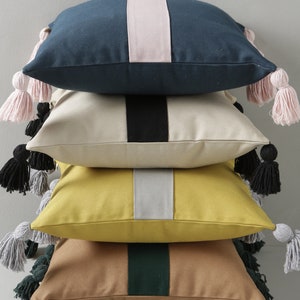 May include: Four decorative throw pillows with different coloured stripes and tassels. The pillows are stacked on top of each other. The top pillow is navy blue with a pink stripe, the second pillow is beige with a black stripe, the third pillow is yellow with a grey stripe, and the bottom pillow is brown with a green stripe.