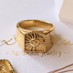 Könnte beinhalten: Goldfarbenes Schmuckset mit einer Anhängerkette und zwei Ringen, alle mit einem Sonnendesign. Der Anhänger hängt an einer filigranen Kette, und die Ringe haben eine quadratische Oberfläche. Der Schmuck wird auf einer weißen Schachtel mit goldenem Text und Weizenähren präsentiert.