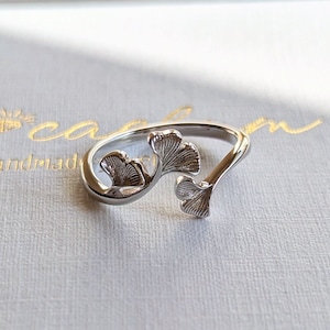 Könnte beinhalten: Ein silberner Ring mit Ginkgoblatt-Design liegt auf einer weißen Schachtel. Der Ring hat zwei detaillierte Ginkgoblätter. Auf der Schachtel steht in Gold "nox carson - handmade". Im Hintergrund ein getrockneter Pflanzenstiel.