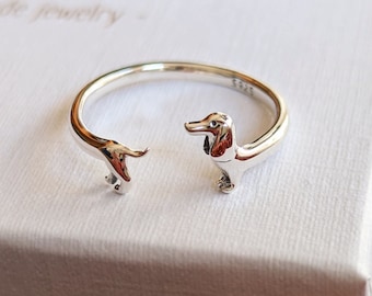 May include: A silver-colored ring featuring a dachshund dog design. The ring has an open design with the dog's head and tail forming the ends. The ring is on a white box with the text "amor caelum handmade jewelry".