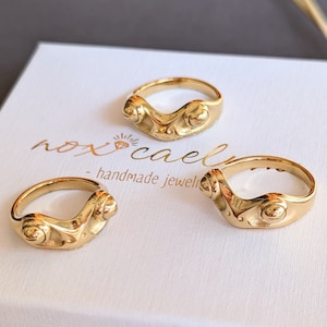 Könnte beinhalten: Drei goldfarbene Ringe mit einem einzigartigen, geformten Design werden auf einer weißen Schachtel präsentiert. Die Ringe haben eine organische Form mit einem zentralen kreisförmigen Element und sind mit "nox caeli - handmade jewel" beschriftet. Ein Weizenähre ist im Hintergrund.