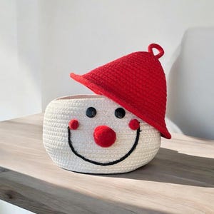 May include: A white woven basket shaped like a snowman's face, with a red hat. The basket has black button eyes, red pom-pom cheeks, and a black stitched smile. The hat has a loop at the top.