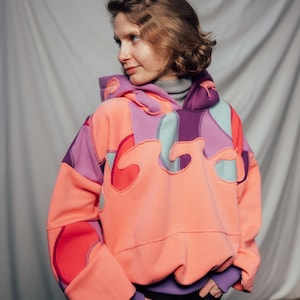 Può includere: Una felpa con cappuccio patchwork colorata nei toni del corallo, viola e verde acqua. La felpa presenta un design unico a forma di fiamma ed è realizzata in tessuto morbido e confortevole. Le maniche e il cappuccio sono accentuati da colori a contrasto.