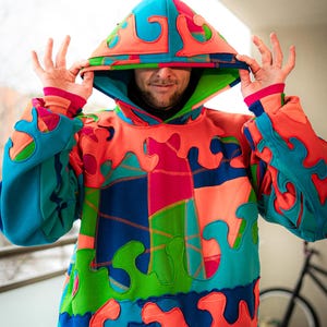 May include: A brightly coloured patchwork hoodie. The design incorporates blue, orange, green, and pink shapes. The hood is up, and the sleeves have matching patterns. The hoodie's style is bold and striking, with a unique, artistic aesthetic.
