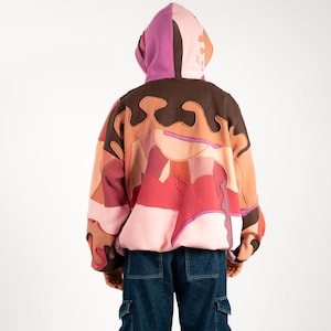 Op de afbeelding: Een patchwork hoodie in roze, bruine en rode tinten, met een bijpassende capuchon. De achterkant toont het abstracte ontwerp en de persoon draagt een donkerblauwe cargobroek.