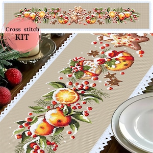 May include: A cross-stitch kit with a festive design. The design includes oranges, berries, gingerbread men, and Christmas ornaments. The text "Cross stitch KIT" is visible. The table runner has a beige background with a white lace trim.