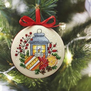 Könnte beinhalten: Ein runder, kreuzgestickter Weihnachtsschmuck mit rotem Band. Der Schmuck zeigt eine Laterne, Beeren und Zitrusfrüchte. Der Hintergrund ist ein verschwommener Weihnachtsbaum.