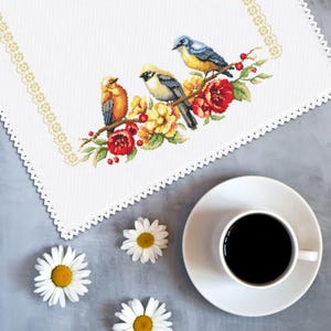May include: A white embroidered table runner with a floral and bird design. The design features three colourful birds perched on a branch with red and yellow flowers. A cup of coffee and daisies are on the table.