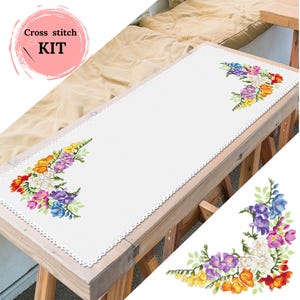 Könnte beinhalten: Weißer Kreuzstich-Tischläufer mit gewelltem Rand und gestickten Blumenmustern in Lila, Gelb, Orange und Blau. Der Text "Cross stitch KIT" befindet sich in einem rosa Kreis oben links.