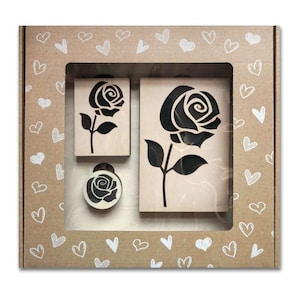 Peut inclure: Un ensemble de trois tampons en bois avec un motif de rose noire. L'ensemble est présenté dans une boîte en carton décorée de coeurs blancs. Il comprend un grand tampon rectangulaire, un petit tampon rectangulaire et un tampon rond.
