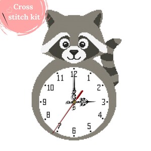 Op de afbeelding: Een grijze kruissteekset met een klok in de vorm van een wasbeer. De wijzerplaat is wit met zwarte cijfers en wijzers, en een rode secondewijzer. De tekst "Cross stitch kit" staat in een roze cirkel.