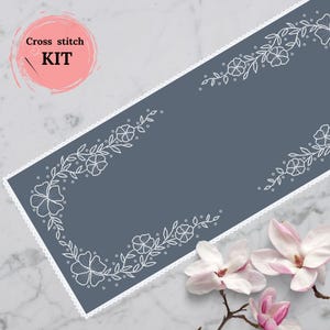 Könnte beinhalten: Ein Kreuzstich-Set mit floralem Design. Das Set enthält ein rechteckiges Stück grauen Stoffes mit weißen gestickten Blumen und Blättern. Die Worte "Cross stitch KIT" befinden sich in einem rosa Kreis.