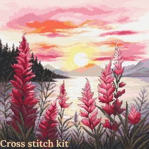 Op de afbeelding: Een kruissteekpakket met een zonsondergangscène boven een meer, met roze en rode bloemen op de voorgrond. De lucht gaat over van oranje naar roze, met een heldere zon. De tekst "Cross stitch kit" staat onderaan.