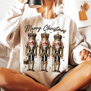 May include: A white sweatshirt with the text "Merry Christmas" and a graphic of three nutcracker figurines wearing gold crowns and leopard print uniforms.