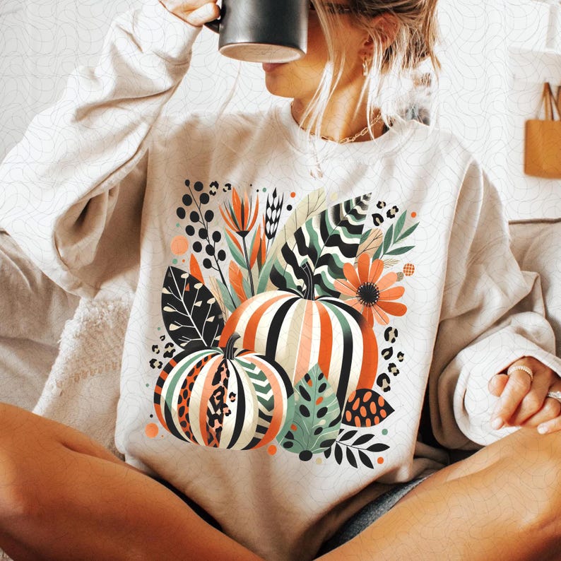 Peut inclure: Un sweat-shirt blanc avec un motif graphique color&eacute; repr&eacute;sentant des citrouilles, des feuilles et des fleurs. Les citrouilles sont ray&eacute;es de noir, de blanc et d'orange. Les feuilles sont vertes et noires. Les fleurs sont orange et rouges.