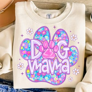 May include: A cream-colored sweatshirt with a purple paw print design. The paw print is filled with a floral pattern in shades of pink, blue, and yellow. The text "Dog Mama" is written in purple letters inside the paw print.