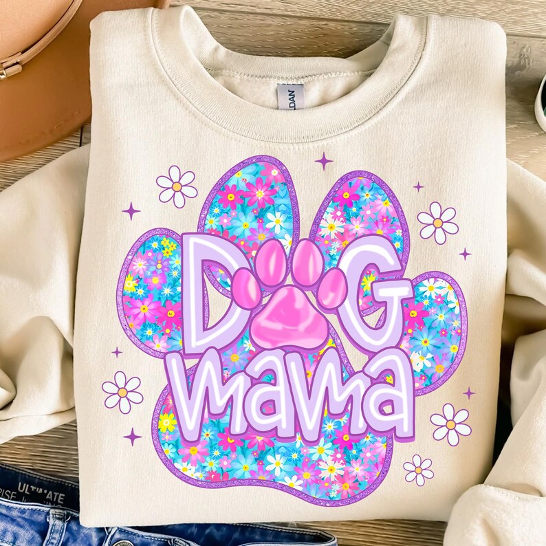 May include: A cream-colored sweatshirt with a purple paw print design. The paw print is filled with a floral pattern in shades of pink, blue, and yellow. The text "Dog Mama" is written in purple letters inside the paw print.