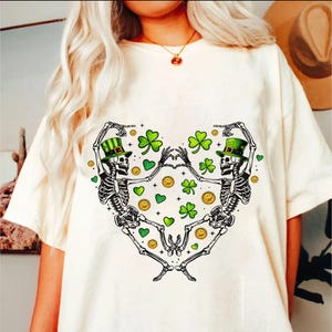 May include: White t-shirt with a graphic of two skeletons wearing green hats and holding hands. The skeletons are surrounded by green shamrocks and gold coins.