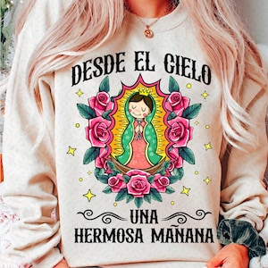 May include: A cream-colored sweatshirt with the text "DESDE EL CIELO UNA HERMOSA MAÑANA" printed on the front. The text is in a black font with a pink and green illustration of a woman in a floral frame.