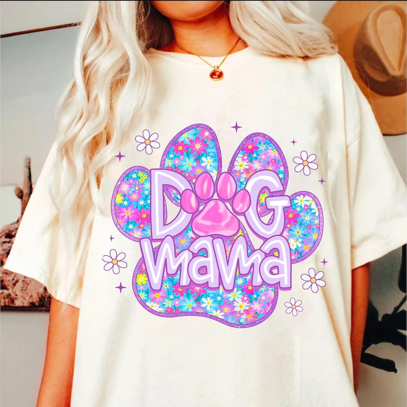 May include: White t-shirt with a purple paw print design. The paw print is filled with colorful flowers and the text "Dog Mama" is written inside the paw print.