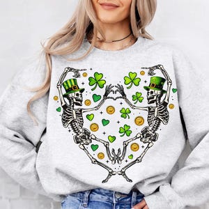 May include: Gray sweatshirt with a graphic of two skeletons wearing green shamrock hats and holding hands. The skeletons are surrounded by shamrocks and gold coins.