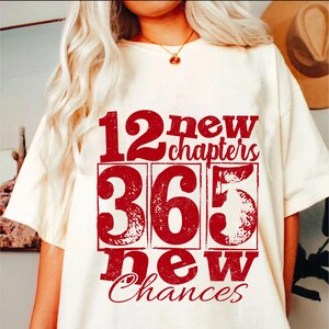 May include: Cream-colored t-shirt with the text "12 new chapters, 365 new chances" in a distressed red font. The design features the numbers 12, 365, and 5 in separate boxes, with the words arranged around them.
