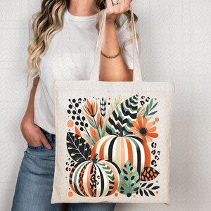 Peut inclure: Un sac fourre-tout blanc avec un motif color&eacute; de citrouilles, de feuilles et de fleurs. Les citrouilles sont ray&eacute;es d'orange et de blanc, et les feuilles sont vertes et noires. Les fleurs sont orange et rouges.