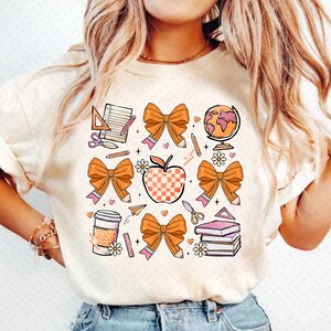May include: A white t-shirt with a design of orange bows, pencils, a red apple, a globe, a coffee cup, scissors, a ruler, a notebook, and books. The design is in a cartoon style.
