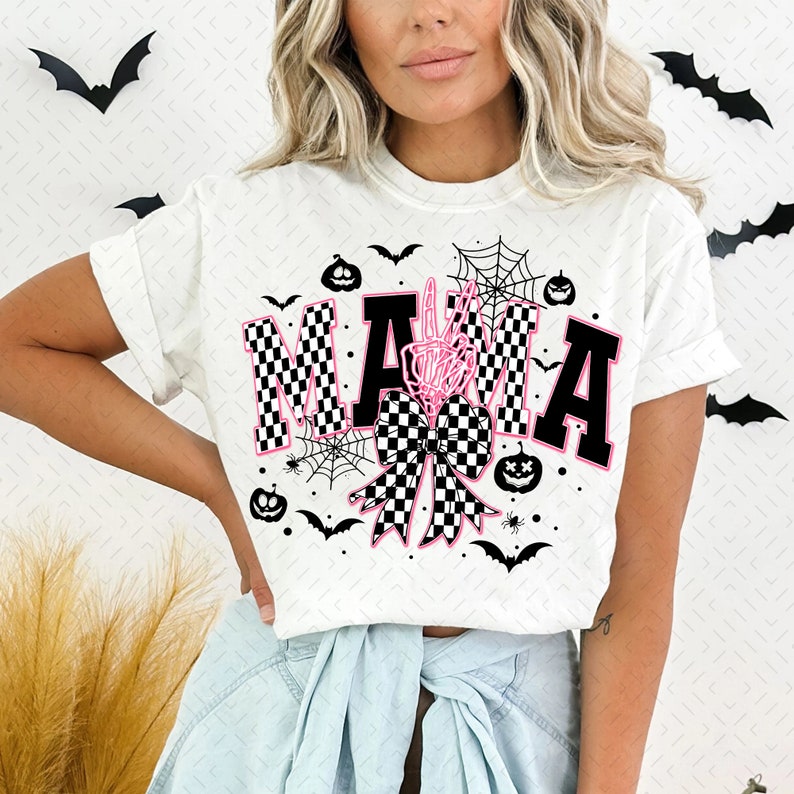 May include: White t-shirt with a black and white checkered "MAMA" graphic with a pink bow and skeleton hand. The graphic also includes bats, spiders, and pumpkins.