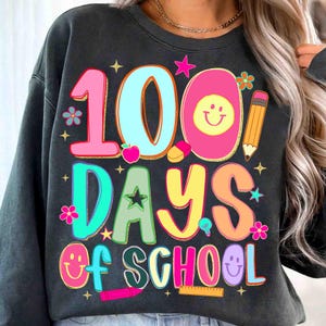 May include: Dark grey sweatshirt with a vibrant graphic celebrating "100 Days of School." The design features the number "100" in pink and blue, and the words "Days of School" in various bright colours. Includes smiley faces, stars, and school-themed illustrations.