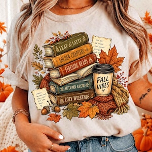 Puede incluir: Una camiseta color crema con un gráfico de una pila de libros con los títulos Leaf & Latte, Golden Chapters, Fireside Reads, Autumn Glow y Cozy Weekends, junto a un vaso de café con la etiqueta Fall Vibes, hojas de otoño y notas de texto.