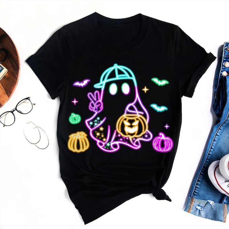 K&ouml;nnte beinhalten: Schwarzes T-Shirt mit einer neon-gr&uuml;nen, blauen und pinkfarbenen Grafik eines Geistes, der einen Hut tr&auml;gt und einen K&uuml;rbis h&auml;lt. Der Geist macht eine Peace-Handgeste. Die Grafik ist von kleineren neon-gr&uuml;nen, blauen und pinkfarbenen K&uuml;rbissen und Flederm&auml;usen umgeben.