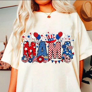 May include: White t-shirt with a red, white, and blue graphic that says "MAMA" with stars, fireworks, and balloons.