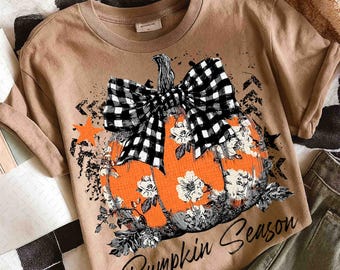 May include: A tan t-shirt featuring a large orange pumpkin design with white floral accents and a black and white checkered bow. The words "Pumpkin Season" are printed in a script font below the pumpkin.