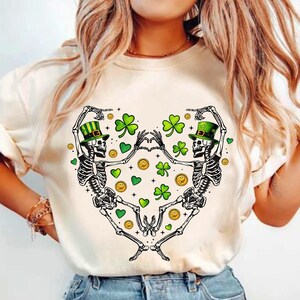 Op de afbeelding: Wit T-shirt met een afbeelding van twee skeletten die groene hoeden dragen en elkaar vasthouden. De skeletten zijn omringd door groene klavers en gouden munten.