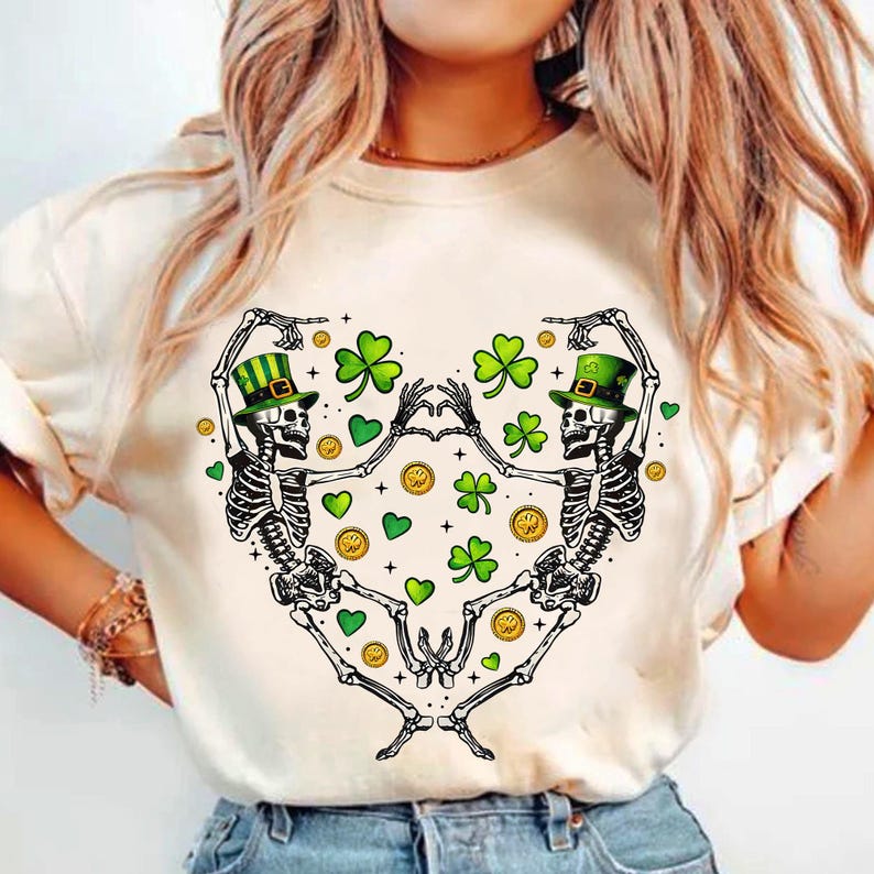 May include: White t-shirt with a graphic of two skeletons wearing green hats and holding hands. The skeletons are surrounded by green shamrocks and gold coins.
