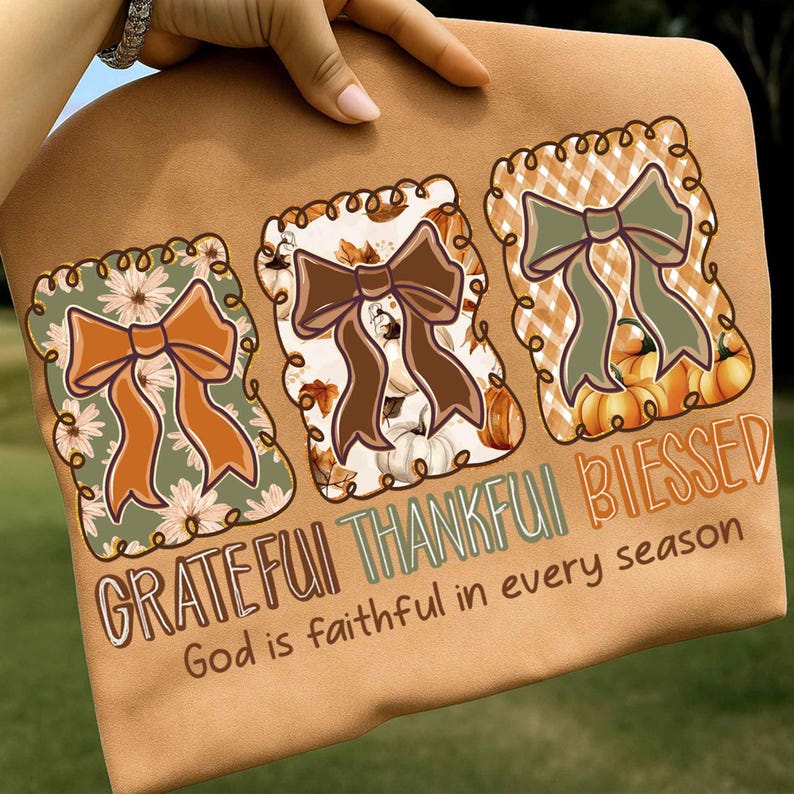 May include: A tan-colored t-shirt featuring three decorative bow designs with the words "Grateful, Thankful, Blessed" and "God is faithful in every season." The bows have fall-themed patterns and colors.