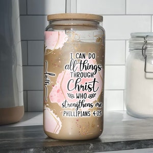 May include: A glass jar candle with a wooden lid. The candle has a gold and pink abstract design with the text "I can do all things through Christ who strengthens me Philippians 4:13".