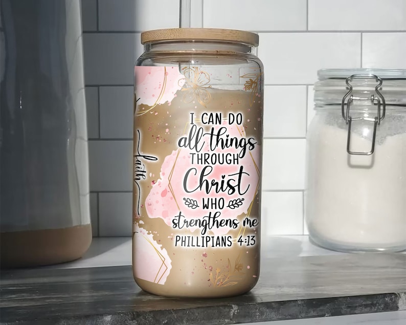 May include: A glass jar candle with a wooden lid. The candle has a gold and pink abstract design with the text "I can do all things through Christ who strengthens me Philippians 4:13".