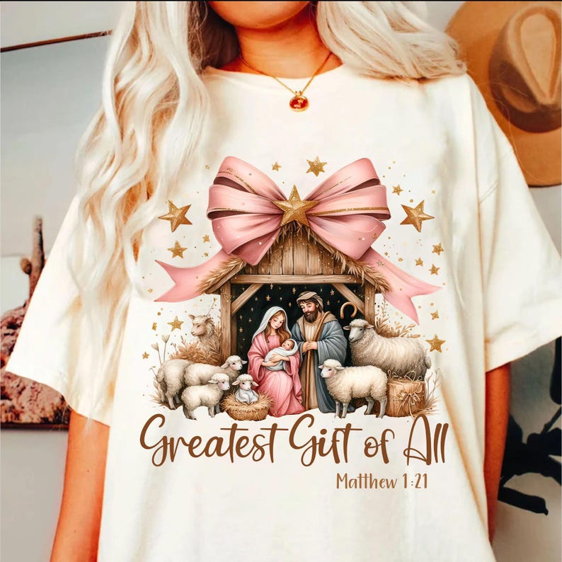 May include: White t-shirt with a pink bow and gold stars. The center of the shirt features a nativity scene with Mary, Joseph, and baby Jesus. The text "Greatest Gift of All" and "Matthew 1:21" is printed below the scene.