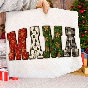 Könnte beinhalten: Ein weißes, weihnachtliches Dekokissen mit dem Wort "MAMA" in großen, festlichen Buchstaben. Die Buchstaben zeigen verschiedene Muster: rote Schneeflocken, grüne Weihnachtsbäume, Lebkuchenmänner und schwarz-weiß kariert.