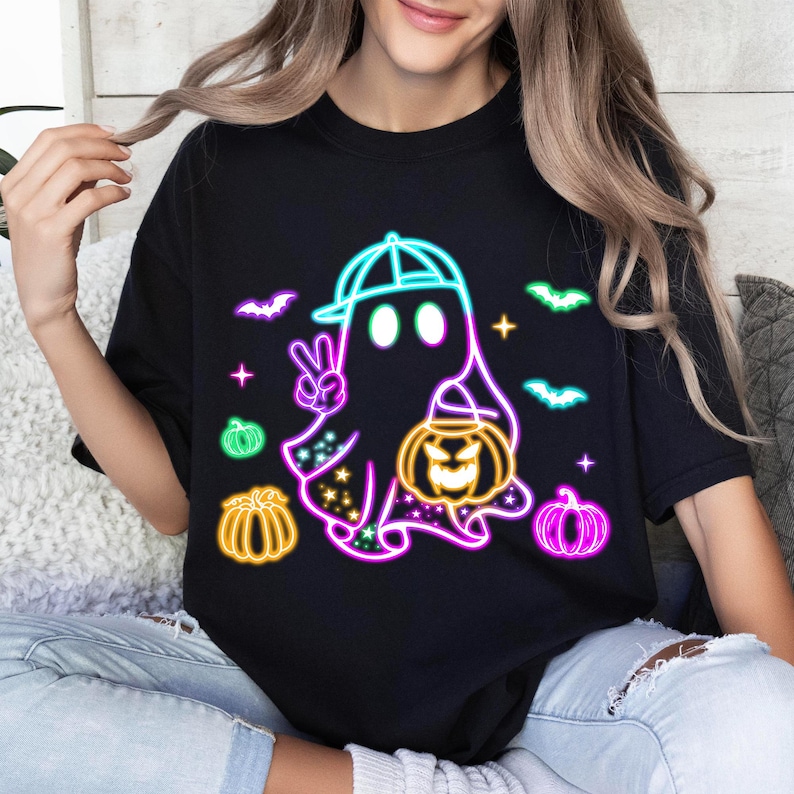 K&ouml;nnte beinhalten: Schwarzes T-Shirt mit einer neon-gr&uuml;nen, blauen und pinkfarbenen Grafik eines Geists, der eine Baseballkappe tr&auml;gt und einen K&uuml;rbis h&auml;lt. Der Geist ist von Flederm&auml;usen und K&uuml;rbissen umgeben.