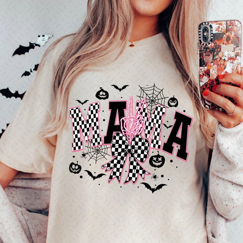 May include: A white t-shirt with a black and white checkered bow and the word "MAMA" in black letters with a pink outline. The letters are decorated with a skeleton hand and spiderwebs. There are also small black bats and pumpkins around the design.