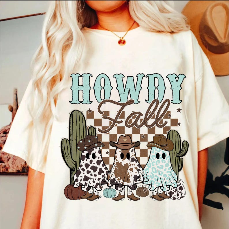 May include: White t-shirt with a graphic design featuring the text "Howdy Fall" in blue and white. The design also includes three ghosts wearing cowboy hats and boots, with a checkered background and cactus plants.