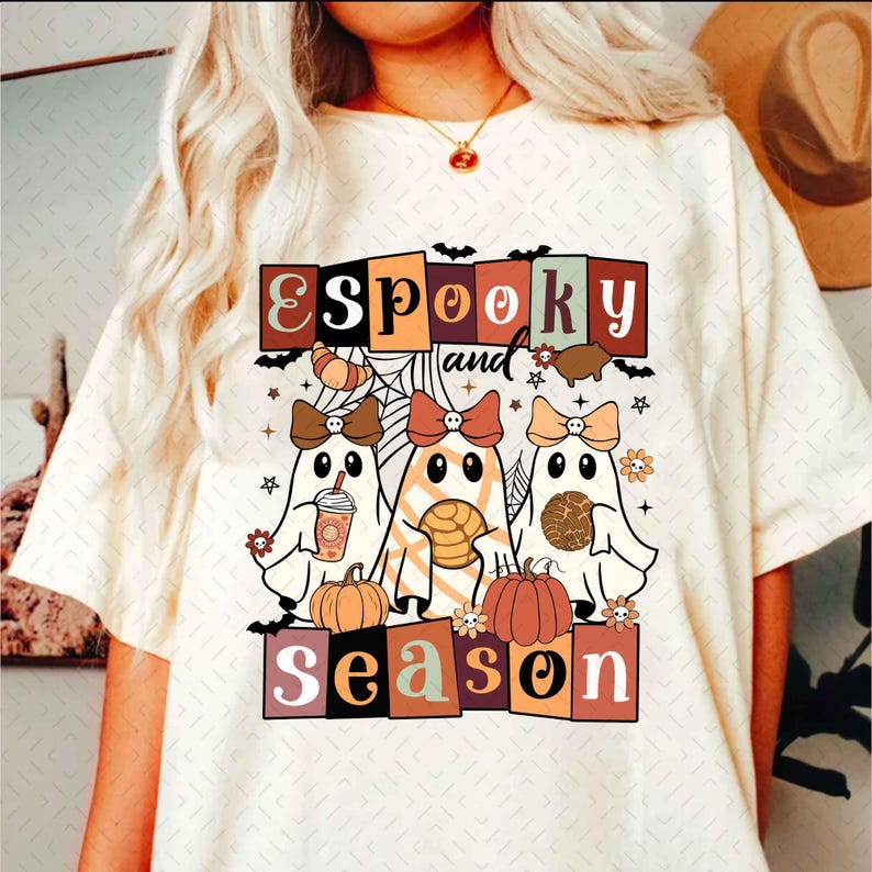 Pu&ograve; includere: Una maglietta bianca con un design grafico che presenta tre fantasmi con fiocchi, che tengono vari dolcetti. Il testo "Spooky and Season" &egrave; scritto in un font retr&ograve; con uno sfondo colorato.