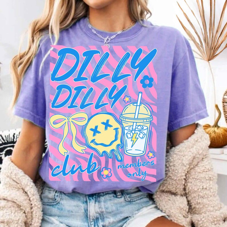 May include: A lilac-colored t-shirt with a graphic design that reads "DILLY DILLY CLUB members only" in blue script. The design includes a yellow bow, a melting yellow smiley face with cross eyes, and a drink with a lightning bolt.