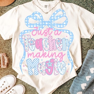 Puede incluir: Camiseta color crema con el texto "Just a Teacher making Magic" en rosa y azul. El diseño incluye un lazo azul a cuadros y estrellas. Se ven un collar y zapatos.