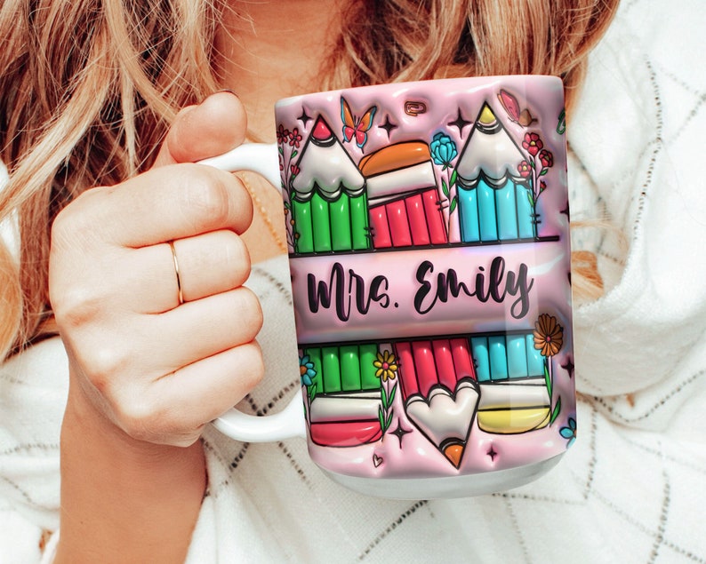 May include: Pink ceramic mug with a colorful design of pencils and flowers. The text "Mrs. Emily" is printed on the mug.